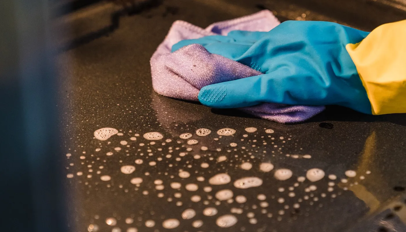 A blue rubber gloved hand wiping a soapy oven surface, the end of tenancy cleaning task that causes the most deposit disputes